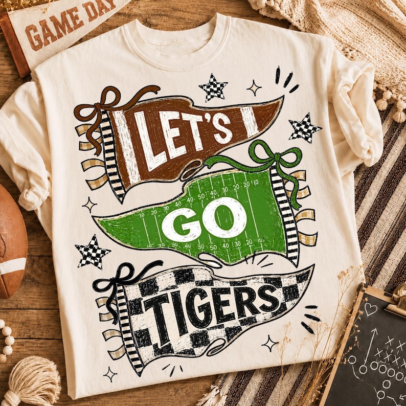 May include: A cream-colored t-shirt featuring a graphic design of three pennants. The top brown pennant says LET'S, the middle green football field pennant says GO, and the bottom checkered pennant says TIGERS. The design includes stars and accents.