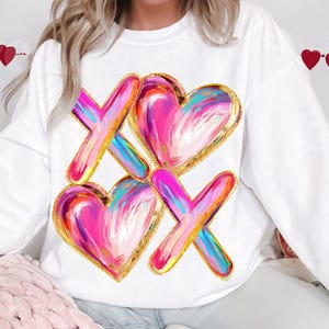 Puede incluir: Sudadera blanca con un dise&ntilde;o de coraz&oacute;n y X de colores. El coraz&oacute;n y la X est&aacute;n pintados en rosa, azul, amarillo y blanco con detalles dorados.