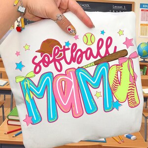 May include: White t-shirt with the words "softball MAMA" in pink and blue, along with a baseball, glove, and stars. The design includes a brown baseball bat and a brown baseball cap.