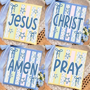 Op de afbeelding: Vier decoratieve vierkante plaquettes met religieuze woorden: "Jezus", "Christus", "Amen" en "Bid". Elke plaquette heeft een kleurrijk patchwork-ontwerp met blauwe en gele accenten, ster- en strikdetails en een blauw lint.