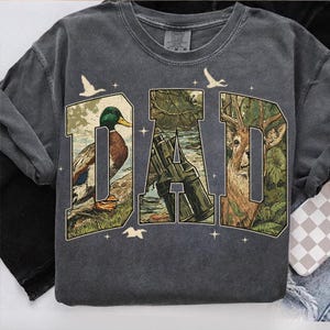 K&ouml;nnte beinhalten: Dunkelgraues Sweatshirt mit dem Wort "DAD" in einem Design zum Thema Natur. Die Buchstaben sind mit Bildern einer Ente, eines Hirsches und eines Jagdgewehrs gef&uuml;llt. Wei&szlig;e V&ouml;gel und Sterne sind um die Buchstaben verstreut.