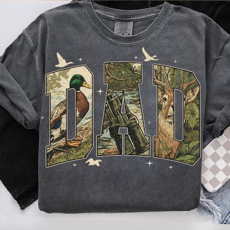K&ouml;nnte beinhalten: Dunkelgraues Sweatshirt mit dem Wort "DAD" in einem Design zum Thema Natur. Die Buchstaben sind mit Bildern einer Ente, eines Hirsches und eines Jagdgewehrs gef&uuml;llt. Wei&szlig;e V&ouml;gel und Sterne sind um die Buchstaben verstreut.
