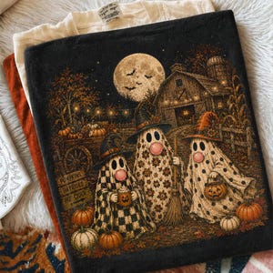 May include: A black t-shirt featuring a graphic illustration of three ghosts wearing witch hats in a farm setting. The scene includes a full moon, a barn, pumpkins, and a sign that reads PUMPKINS, HAYRIDES, HOT CIDER. The ghosts have pink cheeks.