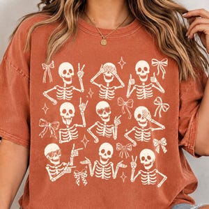 May include: A rust-colored short-sleeve t-shirt featuring a graphic print of nine white skeletons in various poses, including making hand gestures and wearing heart-shaped sunglasses, interspersed with small white bows and four-pointed star icons.