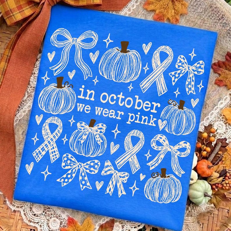 May include: A bright blue fabric featuring a white line-art pattern of pumpkins, ribbons, and bows. The text "in october we wear pink" is printed in the center in a white sans-serif font, surrounded by small heart and star shapes.