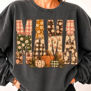 Könnte beinhalten: Ein anthrazitfarbenes Sweatshirt mit Rundhalsausschnitt und dem Wort MAMA in großen Blockbuchstaben. Jeder Buchstabe ist mit einem Patchwork aus Blumen-, Karomustern und Vichy-Karo in Orange, Grün und Weiß gefüllt. Drei Kürbisse liegen am Text.