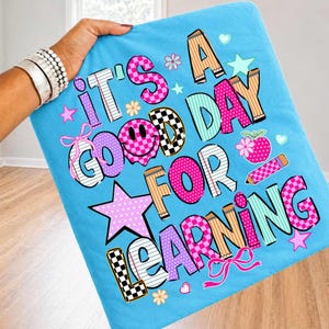 May include: A light blue fabric featuring the text IT'S A GOOD DAY FOR LEARNING in colorful, patterned block letters. The design includes a smiley face, stars, flowers, bows, a pencil, and a red apple graphic in shades of pink, teal, and tan.