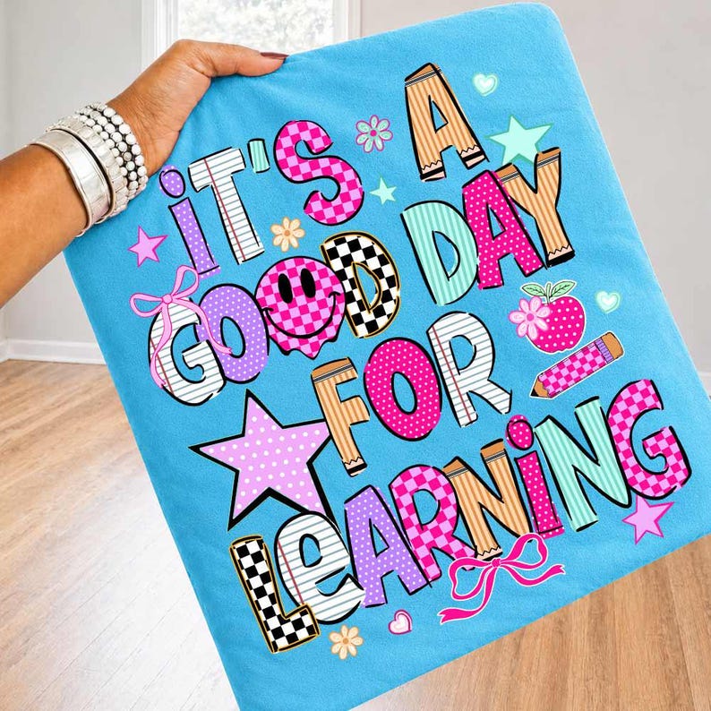 May include: A light blue fabric featuring the text IT'S A GOOD DAY FOR LEARNING in colorful, patterned block letters. The design includes a smiley face, stars, flowers, bows, a pencil, and a red apple graphic in shades of pink, teal, and tan.