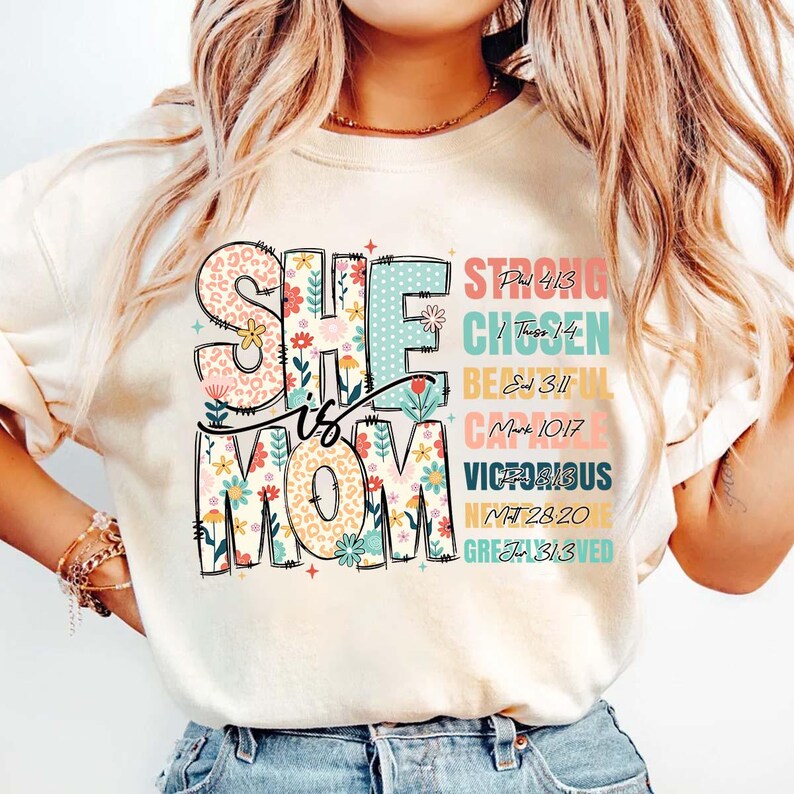 K&ouml;nnte beinhalten: Ein wei&szlig;es T-Shirt mit dem Text "She is Mom" in bunten Buchstaben mit Blumenmustern. Der Text "Strong, Chosen, Beautiful, Capable, Victorious, Greatly Loved" ist rechts neben dem Haupttext vertikal aufgelistet, mit entsprechenden Bibelversen.