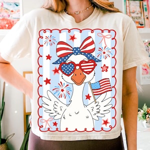 Puede incluir: Camiseta color crema con un dise&ntilde;o patri&oacute;tico de un ganso con gafas de sol en forma de coraz&oacute;n y un lazo, sosteniendo una peque&ntilde;a bandera estadounidense. El dise&ntilde;o incluye elementos rojos, blancos y azules, estrellas y fuegos artificiales.