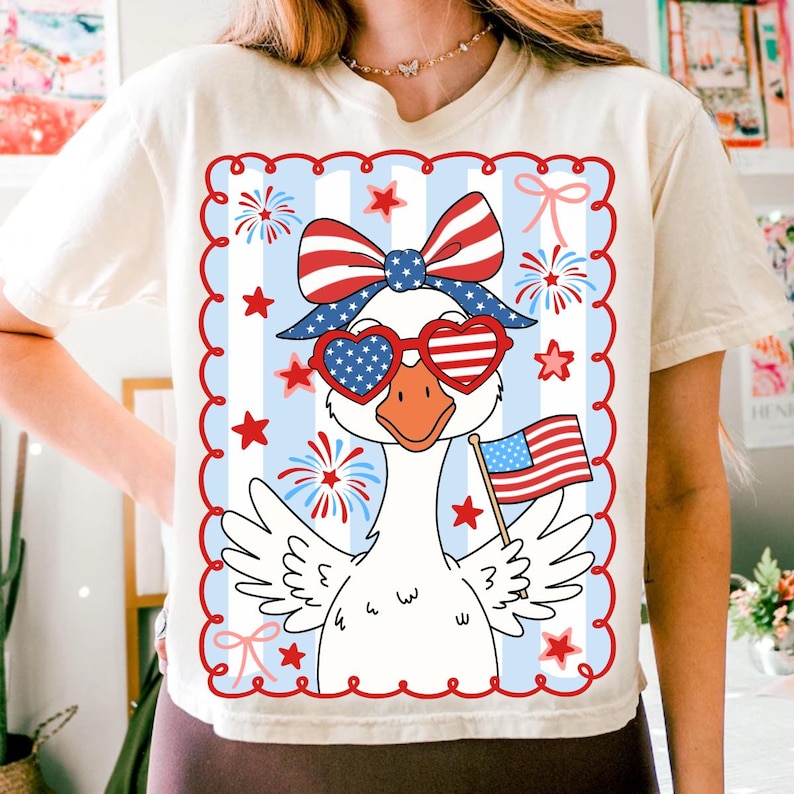 Puede incluir: Camiseta color crema con un dise&ntilde;o patri&oacute;tico de un ganso con gafas de sol en forma de coraz&oacute;n y un lazo, sosteniendo una peque&ntilde;a bandera estadounidense. El dise&ntilde;o incluye elementos rojos, blancos y azules, estrellas y fuegos artificiales.