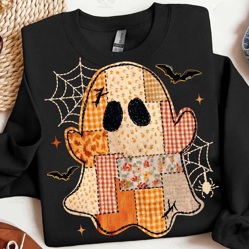 May include: A black crewneck sweatshirt featuring a large central ghost graphic composed of various orange, beige, and white patchwork patterns. The design includes black spiderwebs, small spiders, flying bats, and four-pointed stars surrounding the ghost.