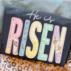May include: A dark grey t-shirt featuring the phrase "He is Risen" in a patchwork style. The letters are in pastel shades with floral and gingham patterns, suitable for Easter.