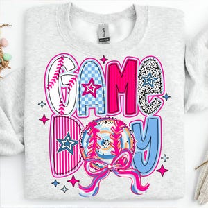 May include: A gray sweatshirt with the words "GAME DAY" in pink, blue, and white letters. The letters are made up of different patterns, including baseball stitching, checkered, and polka dots. A pink and blue baseball with white stitching is in the center of the design.