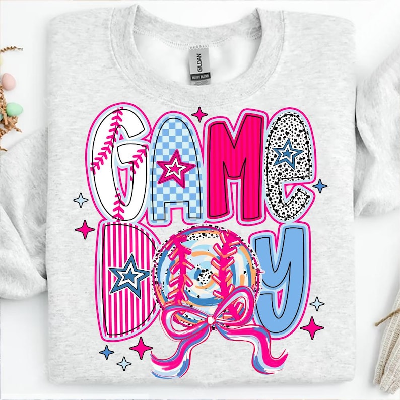 May include: A gray sweatshirt with the words "GAME DAY" in pink, blue, and white letters. The letters are made up of different patterns, including baseball stitching, checkered, and polka dots. A pink and blue baseball with white stitching is in the center of the design.