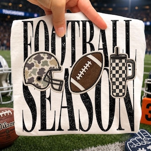 May include: A white t-shirt featuring the black distressed text FOOTBALL SEASON. The design includes three embroidered patches: a camouflage football helmet, a brown football, and a black and white checkered tumbler with a straw.