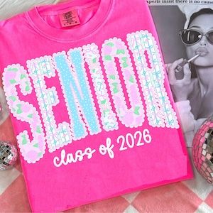 May include: A bright pink t-shirt with the word "SENIOR" in a decorative font, featuring floral and ribbon accents. The text "class of 2026" is printed below. The shirt has a Comfort Colors tag.