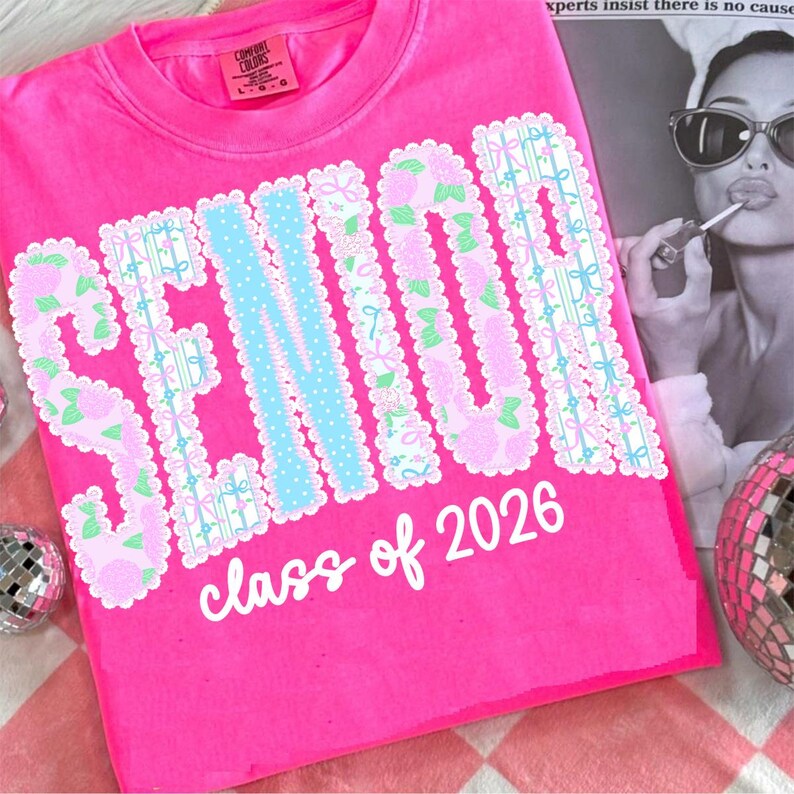 May include: A bright pink t-shirt with the word "SENIOR" in a decorative font, featuring floral and ribbon accents. The text "class of 2026" is printed below. The shirt has a Comfort Colors tag.