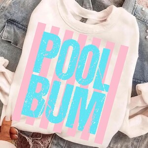May include: A white sweatshirt featuring the words "POOL BUM" in turquoise, distressed lettering, framed by pink vertical stripes. The garment is shown on a denim jacket, giving a relaxed, summery feel.