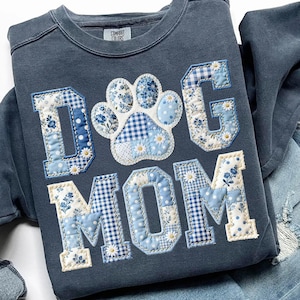 May include: A blue sweatshirt with the words "DOG MOM" in a patchwork design. The letters and paw print are made of various blue and white patterned fabrics, including floral, polka dot, and gingham. The sweatshirt has a crew neck and the brand "Comfort Colors" is visible.