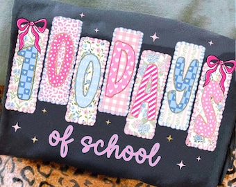 May include: A dark gray t-shirt with the phrase "100 days of school" in pink script. The numbers are in decorative rectangles with floral, animal print, and gingham patterns, and pink bows. Small star accents are scattered around the text.