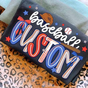 May include: Dark gray t-shirt with a baseball-themed design. The shirt features the words "baseball custom" in a mix of red, white, and blue, along with a baseball, bat, and helmet graphic. Stars and bows add to the design.