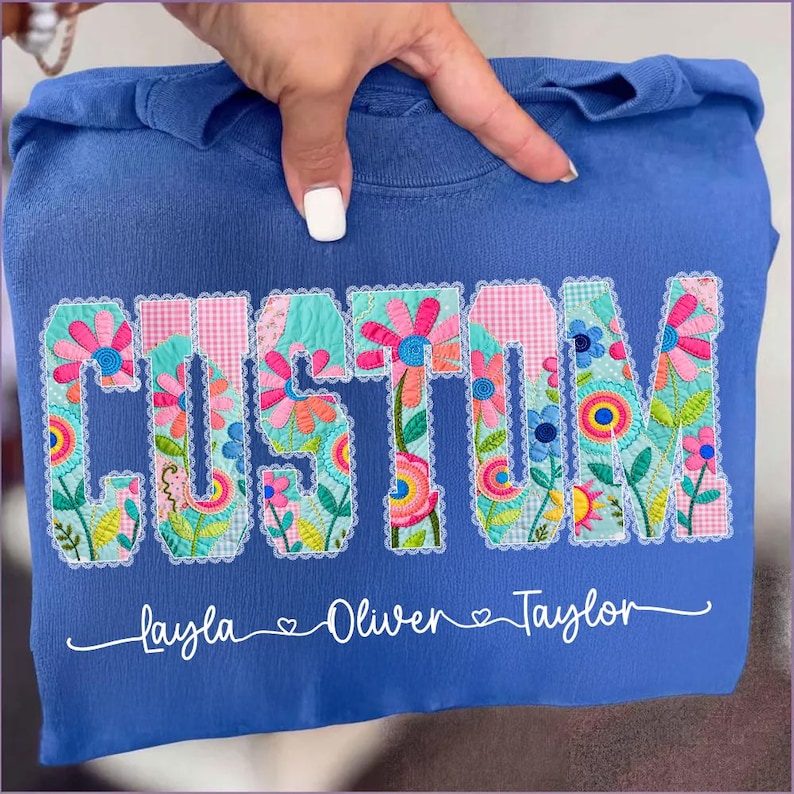 Op de afbeelding: Een blauwe sweatshirt met het woord "CUSTOM" in een bloemenontwerp. Het bloemenontwerp bevat roze, turquoise en gele bloemen. Onder het woord "CUSTOM" staan de namen "layla", "Oliver" en "Taylor".