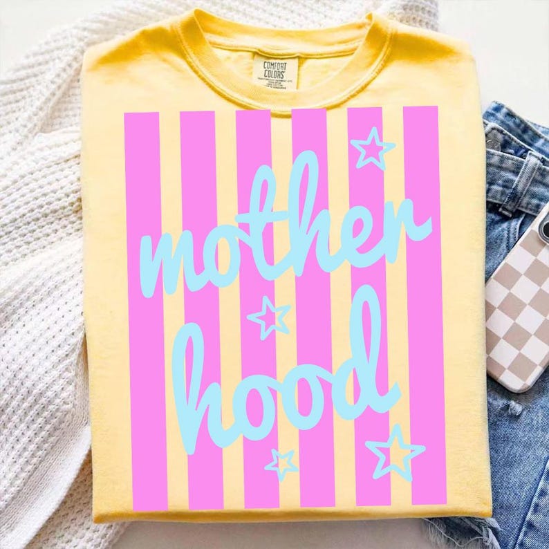 Puede incluir: Camiseta amarilla con rayas verticales rosas y la palabra "motherhood" en escritura azul claro. Estrellas azul claro est&aacute;n dispersas alrededor del texto. La camiseta est&aacute; doblada sobre una superficie blanca con unos vaqueros azules y una funda de tel&eacute;fono.