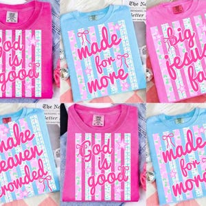 May include: A collection of colorful t-shirts in pink and blue. Each shirt features a unique design with vertical stripes, bows, stars, and text in a contrasting color. The text includes phrases like "God is good" and "Made for more".