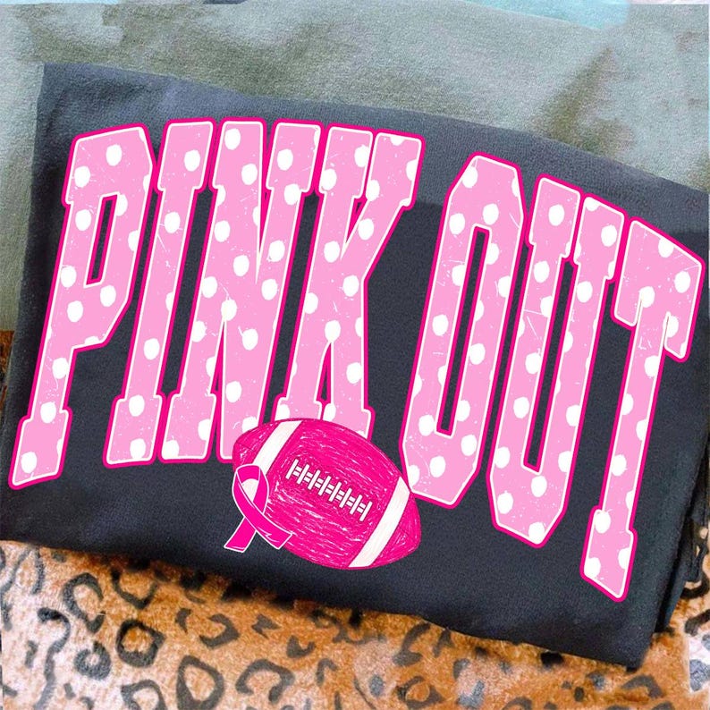 May include: A black t-shirt featuring the text PINK OUT in large, pink, polka-dotted varsity-style letters. Below the text is a pink American football graphic with a pink awareness ribbon integrated into the design of the football.
