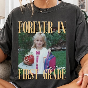 May include: A charcoal gray t-shirt featuring a vintage-style graphic print. The design includes the text FOREVER IN FIRST GRADE, a photograph of a child holding a red gift and a pink bag, and the handwritten-style text Mrs. Taylor.