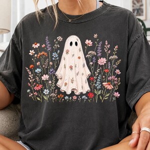 May include: A charcoal gray short-sleeve t-shirt featuring a central graphic of a white ghost covered in small colorful flowers. The ghost is surrounded by a field of various wildflowers in shades of pink, orange, blue, and purple, printed on the front.