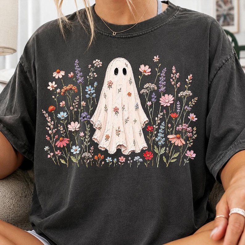 May include: A charcoal gray short-sleeve t-shirt featuring a central graphic of a white ghost covered in small colorful flowers. The ghost is surrounded by a field of various wildflowers in shades of pink, orange, blue, and purple, printed on the front.