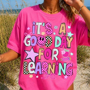 May include: A bright pink short-sleeve t-shirt featuring the text IT'S A GOOD DAY FOR LEARNING in colorful, patterned letters. The design includes a smiley face, stars, hearts, flowers, bows, and pencil graphics in various checkered and polka dot prints.