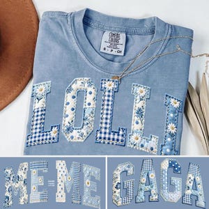 Op de afbeelding: Een lichtblauw Comfort Colors T-shirt met het woord "LOLII" in een patchwork design. De letters hebben bloemen-, stippen- en ruitpatronen in blauwe en witte tinten. Een gouden ketting ligt op het shirt.