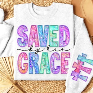 May include: A light gray sweatshirt with the words "SAVED by his GRACE" in colorful, glittery letters. The design includes cross graphics on the sleeve, with a decorative font. The sweatshirt is a casual, comfortable style.