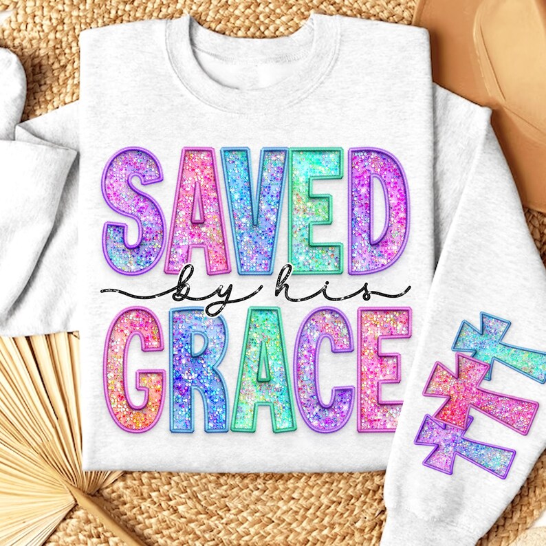 May include: A light gray sweatshirt with the words "SAVED by his GRACE" in colorful, glittery letters. The design includes cross graphics on the sleeve, with a decorative font. The sweatshirt is a casual, comfortable style.