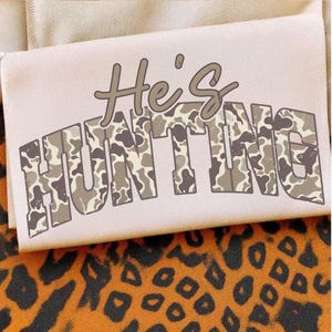 May include: A light pink fabric with the text "He's HUNTING" in a camouflage pattern. The text is in a serif font. The background is a tan fabric and a leopard print fabric.