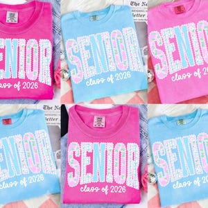 May include: A collection of pink and blue t-shirts. Each shirt features the word "SENIOR" in a decorative font with floral accents, and the text "class of 2026" below. The shirts are displayed on a light-colored surface.