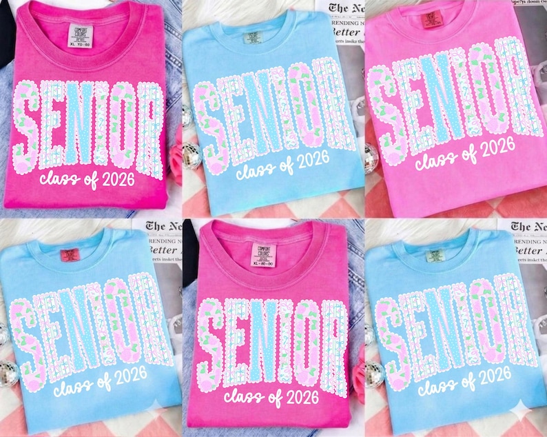 May include: A collection of pink and blue t-shirts. Each shirt features the word "SENIOR" in a decorative font with floral accents, and the text "class of 2026" below. The shirts are displayed on a light-colored surface.