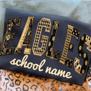 May include: Navy blue sweatshirt with the word "EAGLES" in a patchwork design. The letters feature floral, gingham, and polka dot patterns, with gold accents. The words "school name" are arched below the main text.