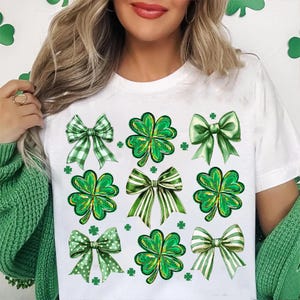 May include: White t-shirt featuring a green shamrock and bow design. The shamrocks and bows are green, white, and yellow.