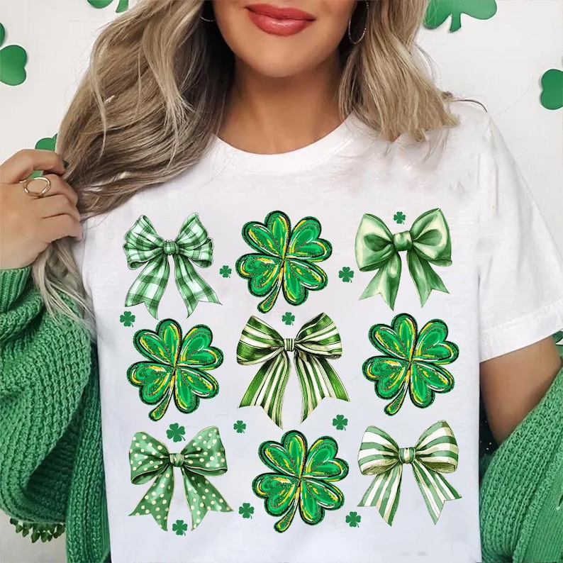 May include: White t-shirt featuring a green shamrock and bow design. The shamrocks and bows are green, white, and yellow.