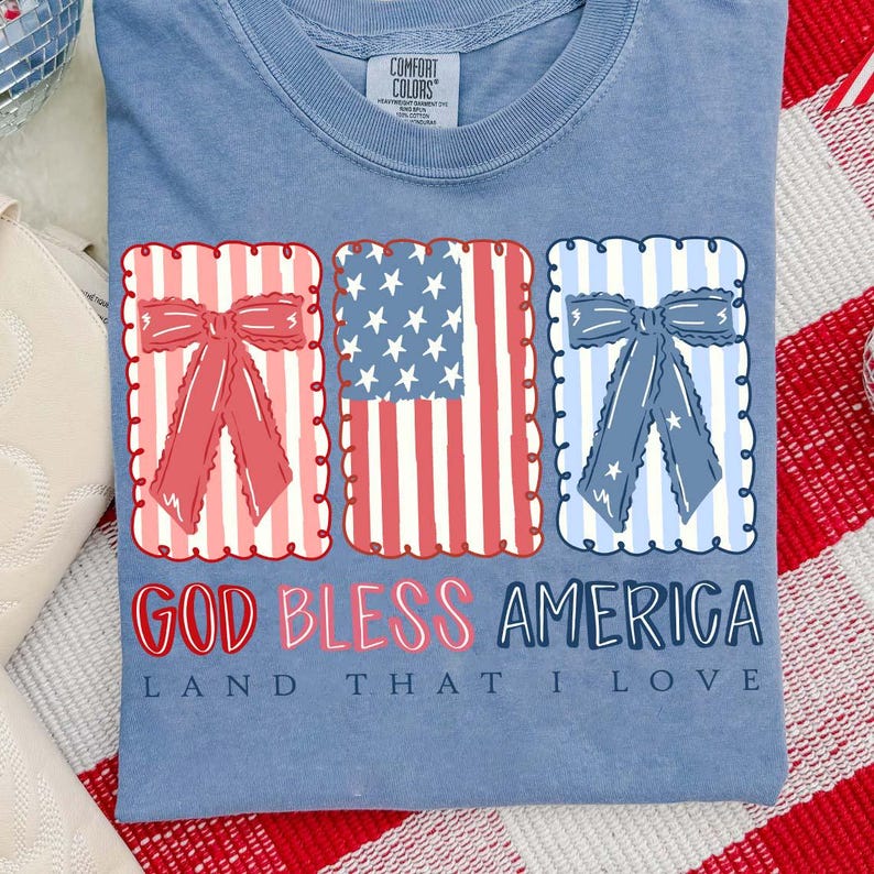 May include: Light blue t-shirt featuring patriotic graphics. The design includes three rectangular frames with bows and American flag elements. The text "GOD BLESS AMERICA LAND THAT I LOVE" is printed below the graphics.