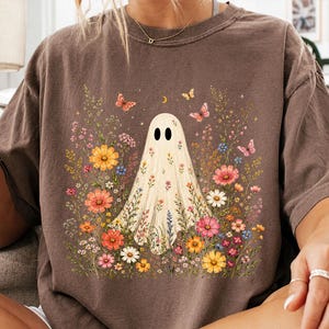 May include: A brown short-sleeve t-shirt featuring a central graphic of a white ghost surrounded by a colorful array of wildflowers, small butterflies, and a crescent moon. The ghost is decorated with a floral pattern across its body.
