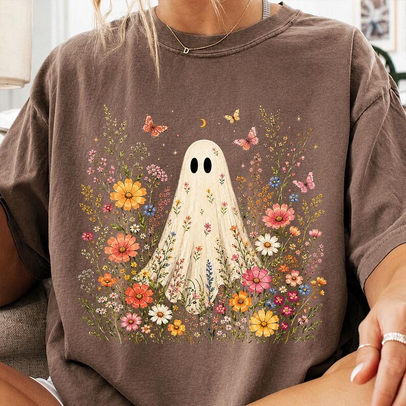 May include: A brown short-sleeve t-shirt featuring a central graphic of a white ghost surrounded by a colorful array of wildflowers, small butterflies, and a crescent moon. The ghost is decorated with a floral pattern across its body.