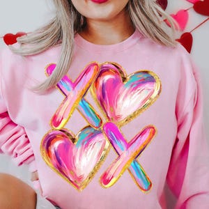 May include: Pink sweatshirt with a graphic design of two hearts and two Xs painted in pink, blue, and gold.