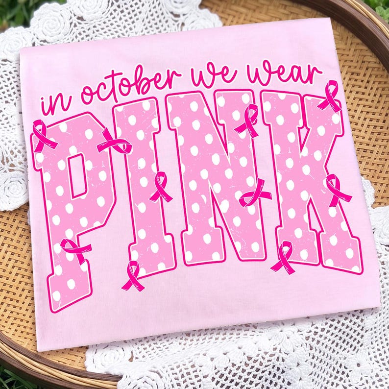 Puede incluir: Una camiseta rosa claro con el texto "in october we wear PINK" en letra cursiva y de molde color rosa oscuro. Las letras grandes de molde tienen un patr&oacute;n de puntos blancos y est&aacute;n decoradas con varios lazos rosas de concienciaci&oacute;n.