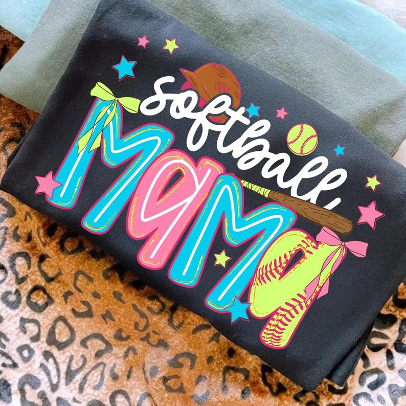 May include: Black t-shirt with the words "softball MOM" in a colorful design. The design includes a softball, glove, bat, and stars. The shirt is folded on top of a leopard print surface.