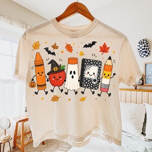 May include: A cream-colored t-shirt featuring a graphic print of five anthropomorphic school supplies and an apple dressed for Halloween. The characters include a crayon, an apple with a witch hat, a ghost, a notebook, and a pencil, surrounded by autumn leaves and bats.